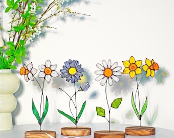 May include: A collection of handcrafted, colorful glass flowers on wooden bases. The flowers include white daisies, yellow daffodils, and a blue flower, each with a unique design. The wooden bases have the word "Cherno" engraved on them.
