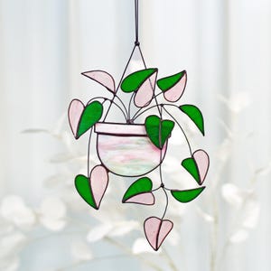 May include: A stained glass hanging of a pink and green heart-shaped plant in a pink pot. The plant is hanging from a black wire.