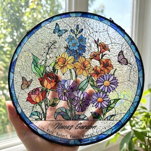 May include: A circular stained glass suncatcher featuring a colorful floral arrangement with roses, daffodils, and daisies, accented by three butterflies. The bottom border includes the text "Nana's Garden" followed by the names Lisa, Kate, Robert, Kevin, George, Eric, and Kris.