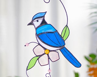 Puede incluir: Un arrendajo azul de vidrio coloreado posado en una flor rosa con hojas verdes. El pájaro tiene un ojo negro y un pico negro. La pieza de vidrio coloreado está colgada de un alambre negro.