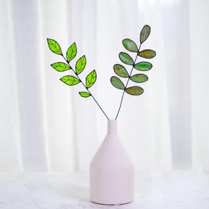 May include: Two green stained glass leaf sprigs in a white ceramic vase. The leaves are different shades of green and have a wire frame.