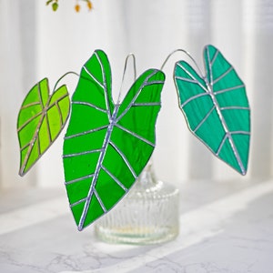 May include: Three stained glass leaves in shades of green, mounted on a wire stem, and displayed in a clear glass vase. The leaves are shaped like the leaves of a tropical plant.