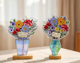 May include: Two stained glass floral arrangements in vases on wooden bases. Each features colorful flowers and a rectangular label reading "Nana's Garden" with names listed below. The left vase is iridescent blue, and the right is green.