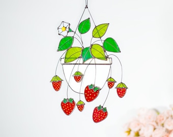 May include: A stained glass hanging strawberry plant with red berries, green leaves, and a white flower with a yellow center. The plant is suspended by a black wire. The berries have a speckled pattern.