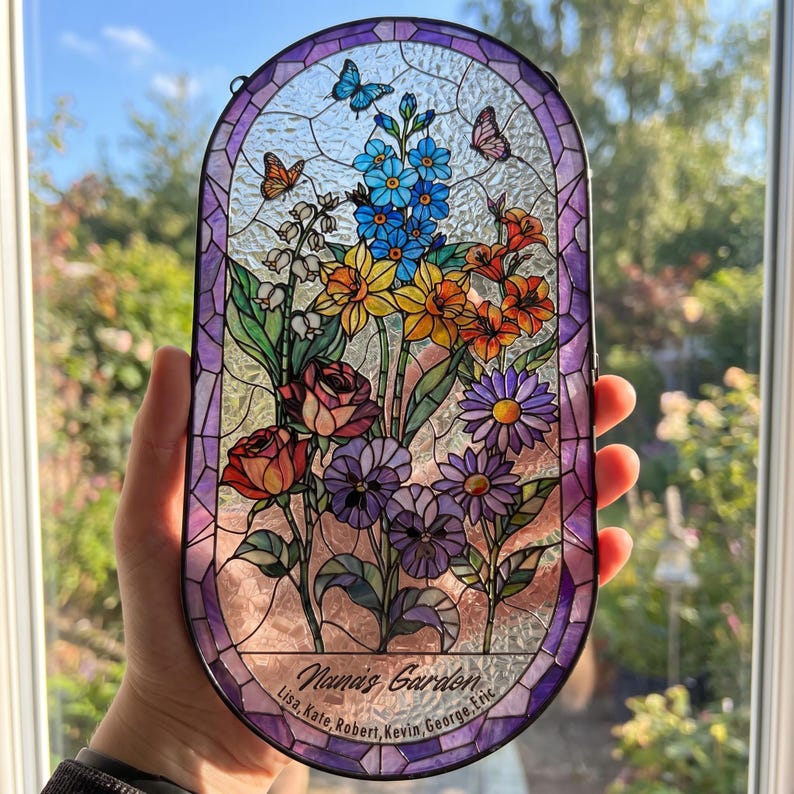 May include: An oval stained glass suncatcher featuring a floral arrangement of blue, yellow, orange, red, and purple flowers with butterflies. The glass has a purple border and includes the text "Nana's Garden" with the names Lisa, Kate, Robert, Kevin, George, Eric.