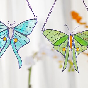 Puede incluir: Dos atrapasoles de vidrio coloreado en forma de mariposa colgando de cadenas. La mariposa de la izquierda es azul y verde con detalles amarillos. La mariposa de la derecha es verde con detalles naranjas.