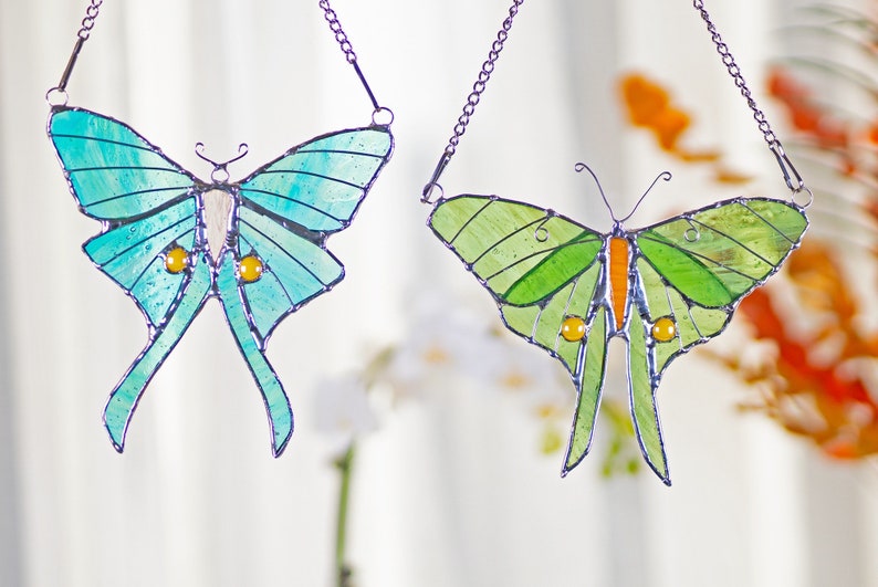 Puede incluir: Dos atrapasoles de vidrio coloreado en forma de mariposa colgando de cadenas. La mariposa de la izquierda es azul y verde con detalles amarillos. La mariposa de la derecha es verde con detalles naranjas.
