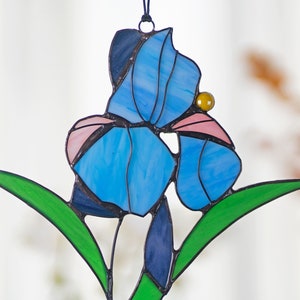 May include: A stained glass suncatcher featuring a blue iris flower with pink accents and a yellow bead. The flower is surrounded by green leaves.