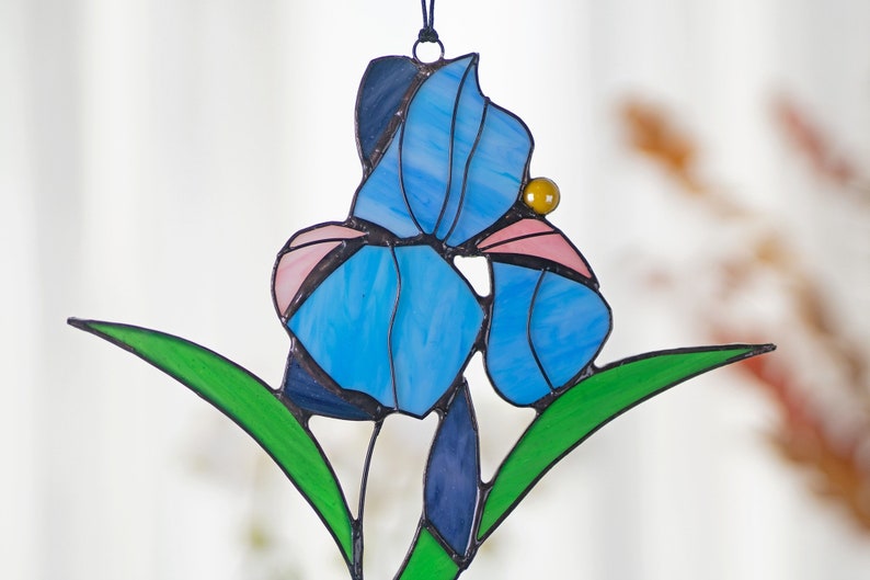 May include: A stained glass suncatcher featuring a blue iris flower with pink accents and a yellow bead. The flower is surrounded by green leaves.