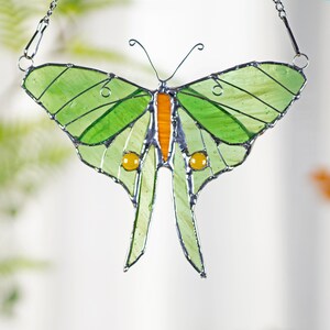 Puede incluir: Un atrapasol de vidrio coloreado en forma de mariposa con alas verdes, acentos naranja y amarillos y una cadena plateada para colgar.