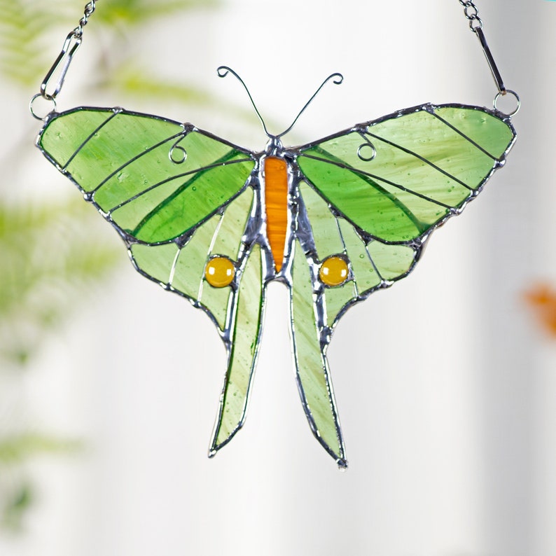 Puede incluir: Un atrapasol de vidrio coloreado en forma de mariposa con alas verdes, acentos naranja y amarillos y una cadena plateada para colgar.