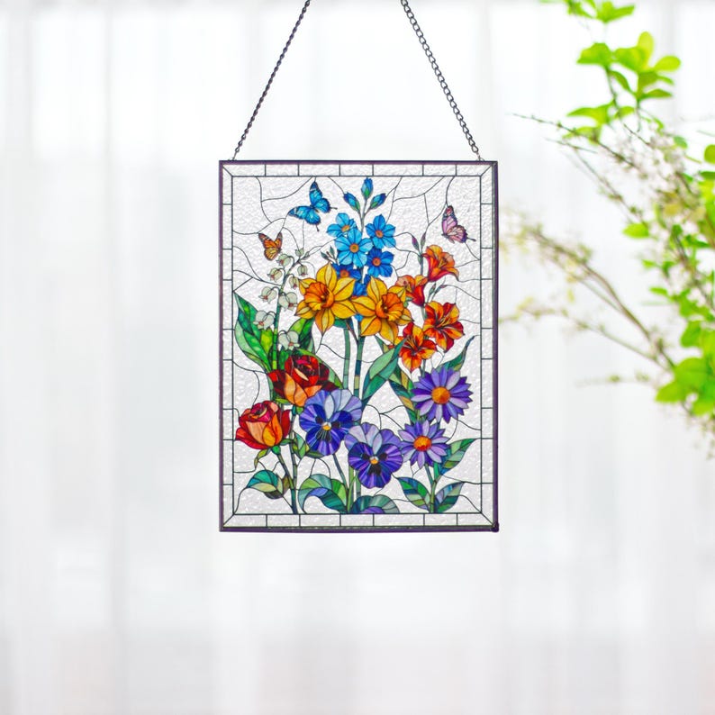 May include: A rectangular stained glass suncatcher featuring a vibrant floral arrangement with red roses, yellow daffodils, blue forget-me-nots, purple pansies, and orange lilies, accented by two small butterflies, all enclosed in a dark metal frame.