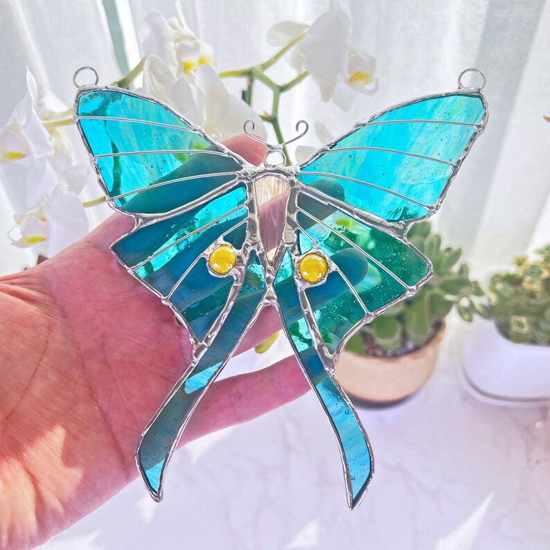 Puede incluir: Una mariposa de vidrio emplomado con alas de color verde azulado y detalles amarillos. La mariposa est&aacute; colgada de una cuerda y est&aacute; enfocada.