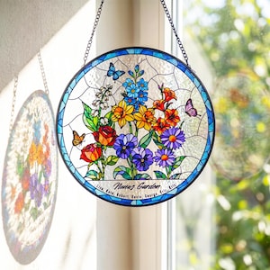 May include: A circular stained glass suncatcher featuring a vibrant floral arrangement with red roses, yellow daffodils, blue flowers, and purple daisies. Butterflies are interspersed among the blooms. The text reads Nana's Garden with names below.