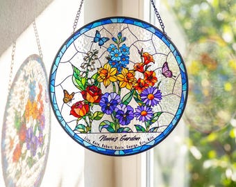 May include: A circular stained glass suncatcher featuring a vibrant floral arrangement with red roses, yellow daffodils, blue flowers, and purple daisies. Butterflies are interspersed among the blooms. The text reads Nana's Garden with names below.