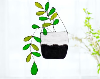 May include: Stained glass hanging planter with green and yellow leaves. The planter is black and white, with a wavy line separating the colors. The leaves are a mix of green and yellow, and the piece is suspended by a thin wire.