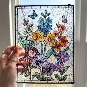 May include: A rectangular stained glass panel featuring a vibrant floral arrangement with yellow daffodils, pink roses, purple pansies, blue forget-me-nots, and orange lilies. Two butterflies are depicted among the flowers and green leaves.
