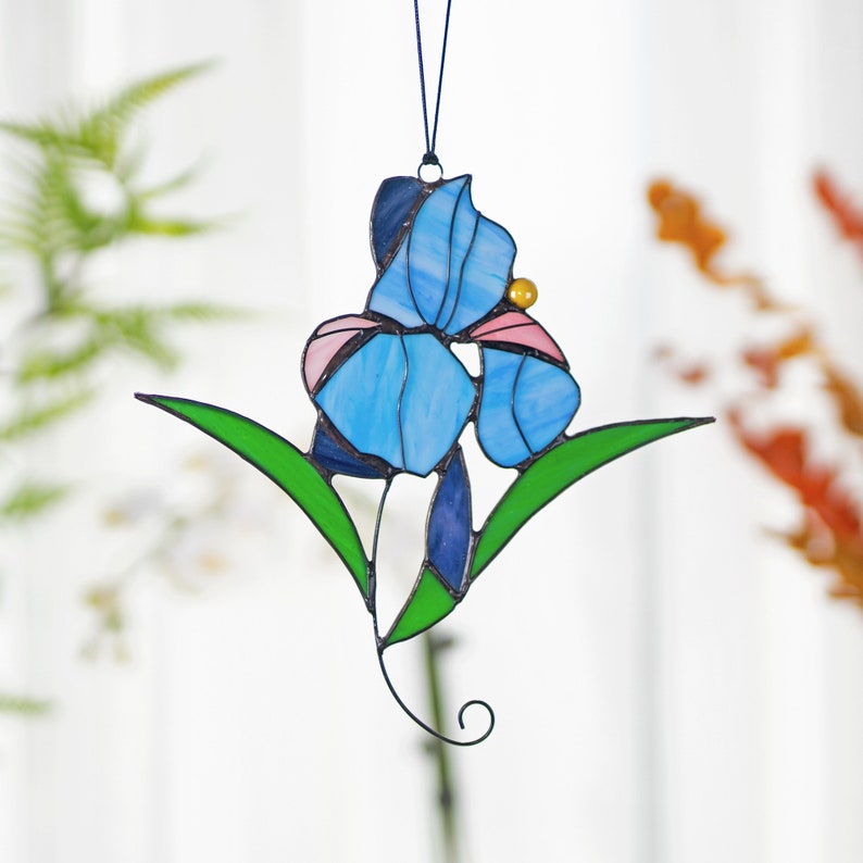 May include: A stained glass window hanging with a blue and pink iris flower with green leaves.