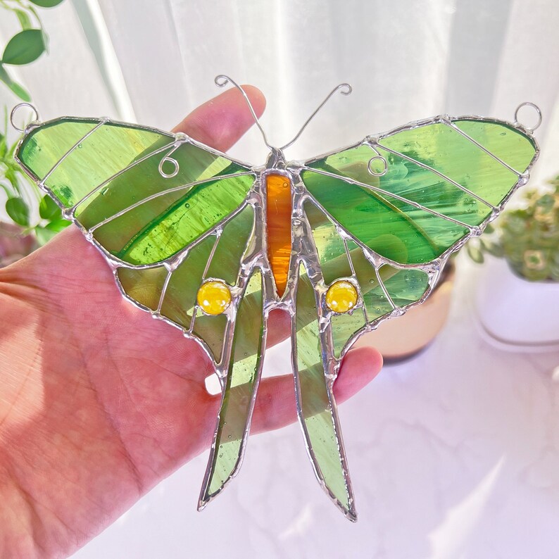 Puede incluir: Una mariposa de vidrio emplomado con alas verdes y detalles amarillos. La mariposa est&aacute; hecha de vidrio y tiene un marco de alambre.