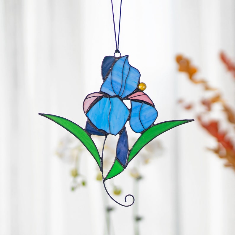 May include: A stained glass hanging featuring a blue iris flower with pink accents and green leaves. The flower is suspended from a black string.