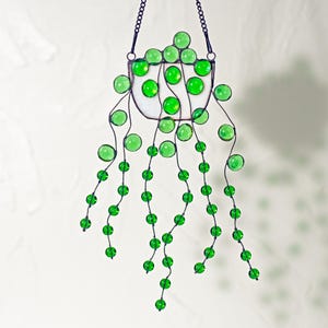 Op de afbeelding: Een gebrandschilderd zonnenvanger met een wit, halvemaanvormig frame en groene glazen kralen. De kralen zijn bovenaan in een tros gerangschikt en hangen in strengen naar beneden.