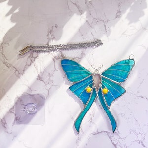 Puede incluir: Un atrapasol de vidrio coloreado en forma de mariposa con un dise&ntilde;o iridiscente azul y verde. La mariposa tiene detalles amarillos y est&aacute; colgada de una cadena plateada.