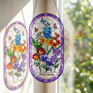 May include: An oval stained glass suncatcher featuring a floral arrangement with blue, yellow, orange, red, and purple flowers and butterflies. The text "Nana's Garden" and names "Lisa, Kate, Robert, Kevin, George, Eric" are printed at the bottom.