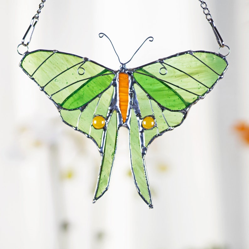 Puede incluir: Un atrapasol de vidrio coloreado en forma de mariposa con alas verdes, acentos naranja y amarillos y una cadena plateada.