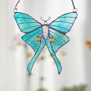 Puede incluir: Una ventana de vitral colgante con una polilla luna azul turquesa con ojos amarillos. La polilla est&aacute; suspendida de una cadena plateada.