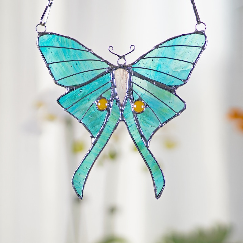 Puede incluir: Una ventana de vitral colgante con una polilla luna azul turquesa con ojos amarillos. La polilla est&aacute; suspendida de una cadena plateada.