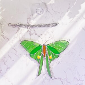Puede incluir: Una mariposa de vidrio coloreado con alas verdes, detalles naranjas y amarillos, y una cadena plateada para colgar.