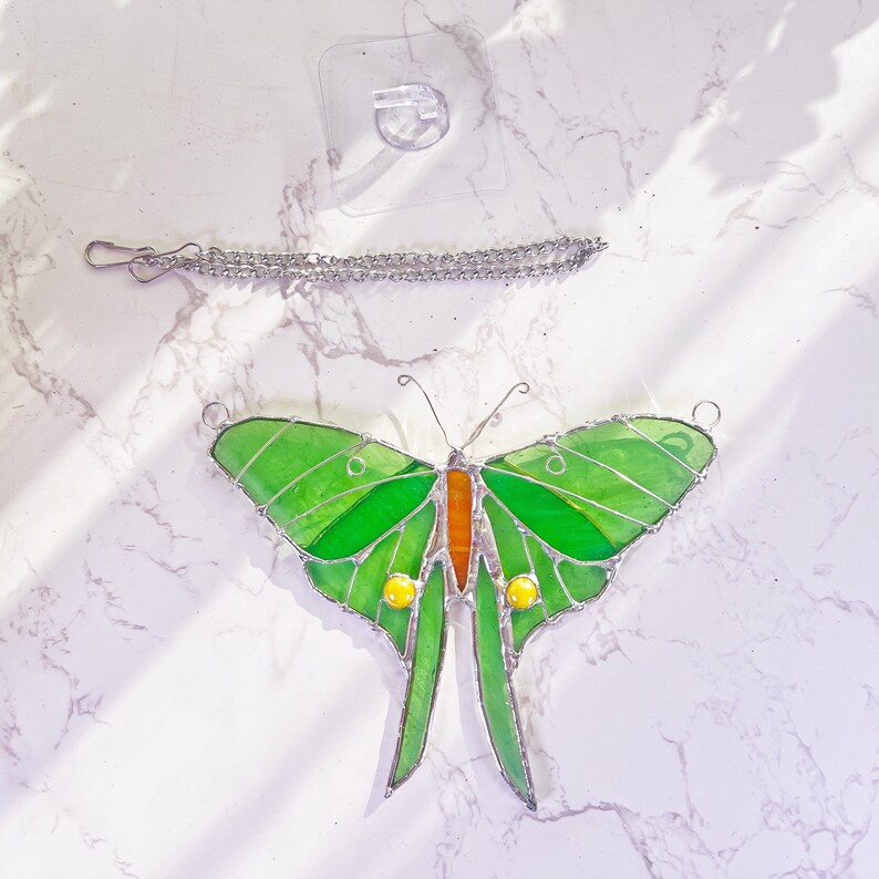 Puede incluir: Una mariposa de vidrio coloreado con alas verdes, detalles naranjas y amarillos, y una cadena plateada para colgar.