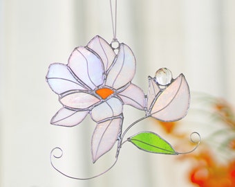 May include: A stained glass flower suncatcher with a white and iridescent flower, a green leaf, and a clear glass bead. The suncatcher is hanging from a white string.