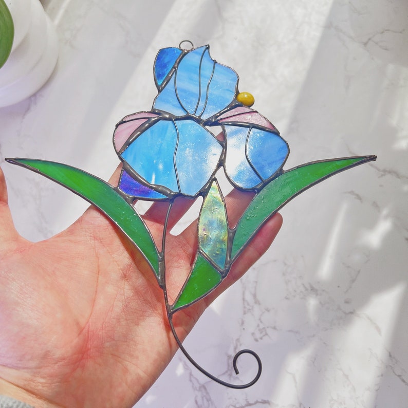May include: A stained glass window hanging features a blue and pink iris flower with green leaves. The flower has a yellow center.