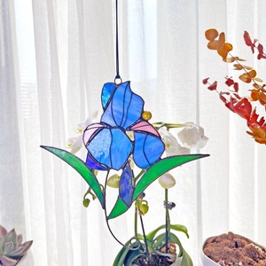 May include: A stained glass window hanging features a blue and pink iris flower with green leaves. The flower is suspended from a black string.