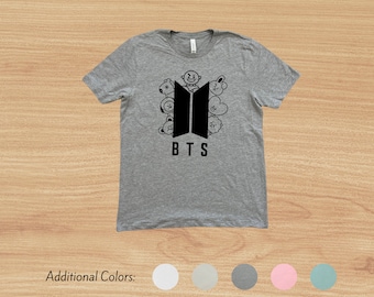 May include: Gray t-shirt with a black BTS logo and cartoon characters surrounding it. The text "BTS" is below the logo.