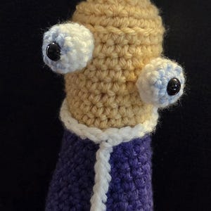 Puede incluir: Muñeca de ganchillo hecha a mano con cabeza beige, ojos grandes blancos y azules y cuerpo morado. La muñeca tiene un ribete blanco y una corona en forma de estrella. Una creación única y caprichosa.