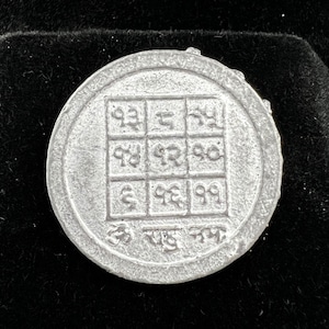 May include: A silver-colored metal charm with a square grid pattern and numbers inscribed inside. The charm is in a black velvet box.