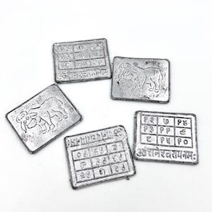 Könnte beinhalten: Fünf silberne Metall-Anhänger mit verschiedenen Designs. Ein Anhänger zeigt einen Stier, ein anderer ein quadratisches Gitter mit Zahlen, und die restlichen drei ein quadratisches Gitter mit Zahlen und Text in einer Fremdsprache.