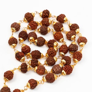 Puede incluir: Un collar mala marrón y dorado con cuentas hechas de semillas de rudraksha. El collar está encordado con metal de color dorado.