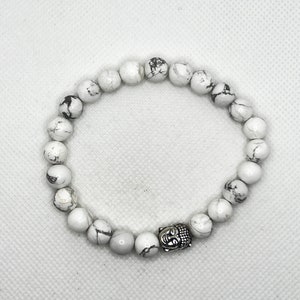 May include: White Howlite beaded bracelet with a silver Buddha charm. The beads are round and have a marbled pattern.