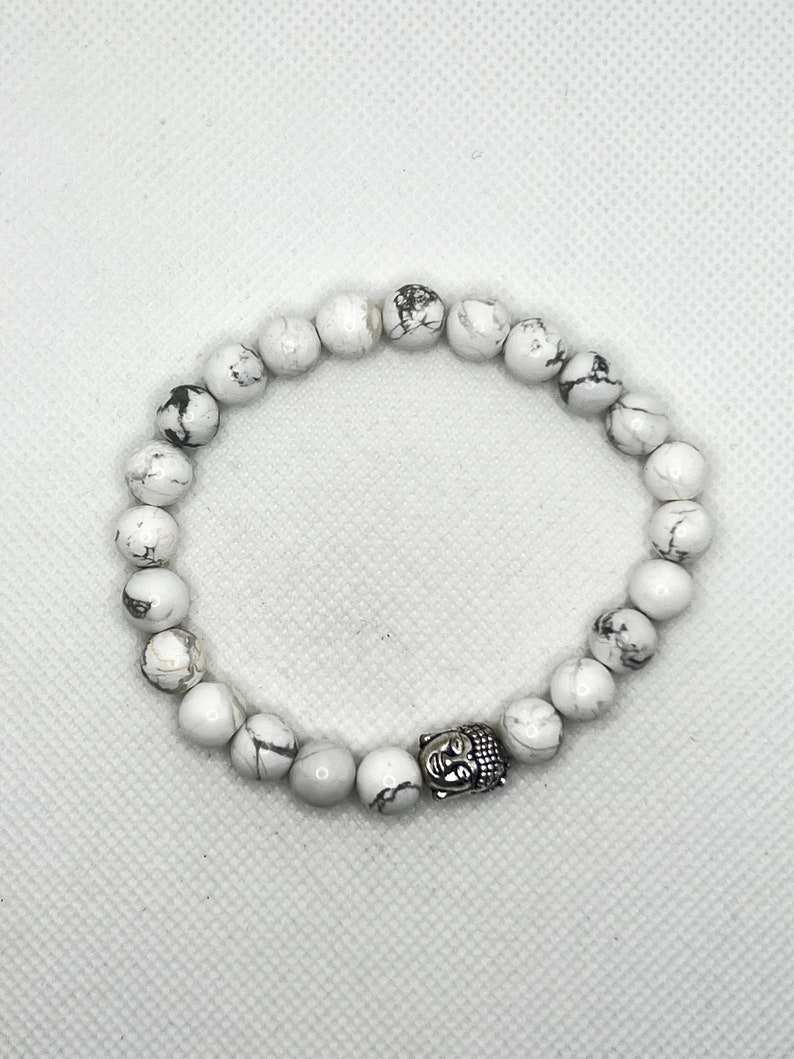 May include: White Howlite beaded bracelet with a silver Buddha charm. The beads are round and have a marbled pattern.
