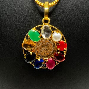 Puede incluir: Un collar de oro con un colgante circular que presenta nueve piedras preciosas de varios colores, incluyendo rojo, verde, azul y blanco. El centro del colgante presenta un diseño dorado con detalles intrincados.