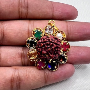 Puede incluir: Un colgante de oro con una cuenta de rudraksha roja en el centro rodeada por un círculo de piedras preciosas de colores. Las piedras preciosas están engastadas en oro e incluyen rojo, azul, verde, amarillo y blanco.