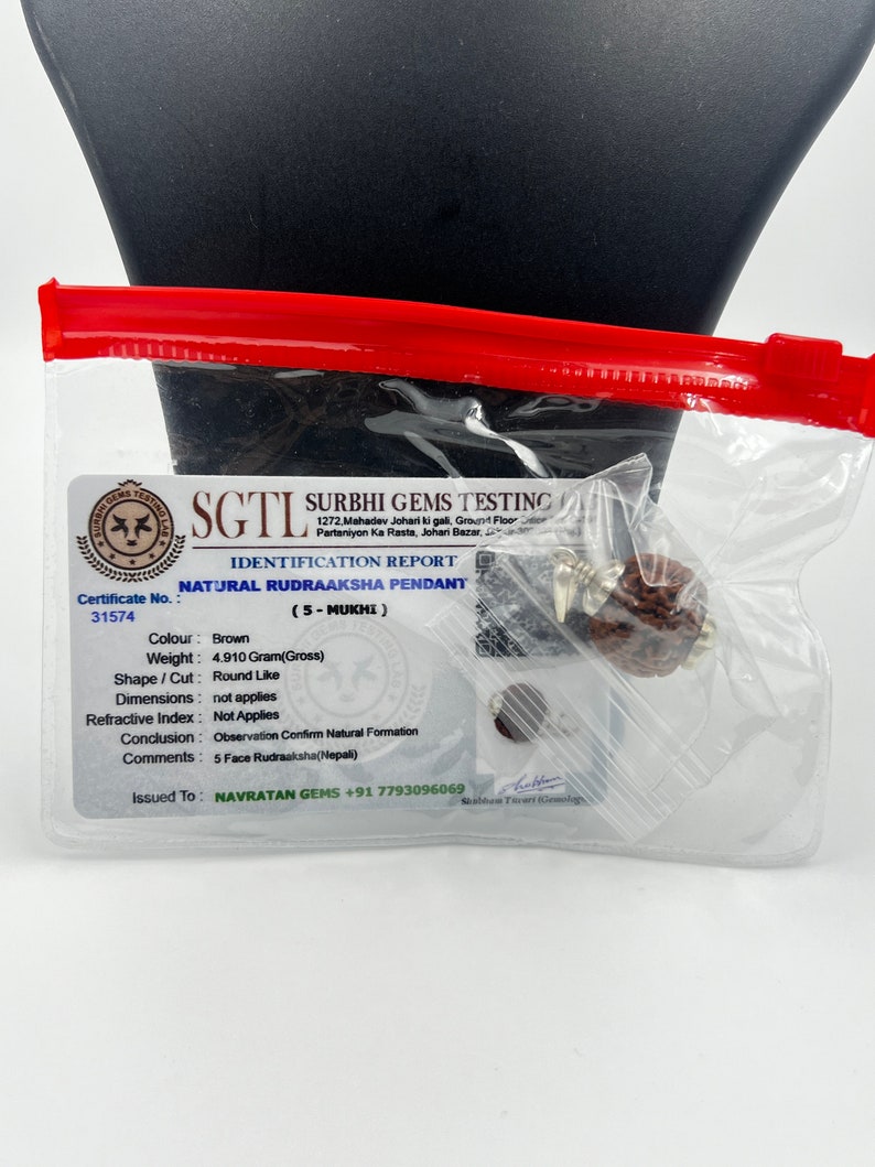 May include: A brown Rudraksha pendant with a 5-Mukhi certification card. The card includes details like weight, shape, and origin. The card is issued by Surbhi Gems Testing.