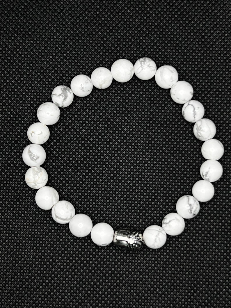 May include: White gemstone bracelet with a silver charm. The bracelet is made of round white beads and has a silver charm with a design on it.