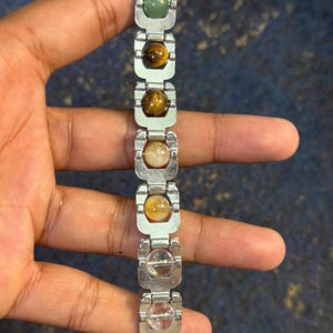 Könnte beinhalten: Ein silberfarbenes Armband mit einer Reihe von polierten, farbigen Edelsteinen. Das Armband enthält grüne, braune, gelbe und klare Steine, die durch rechteckige Metallglieder verbunden sind. Das Armband ist etwa 18 cm lang.