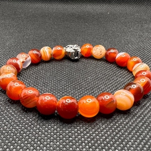 May include: A red carnelian gemstone bracelet with a silver clasp. The beads are round and have a striped pattern.