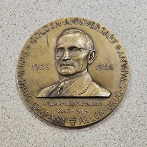 May include: A bronze commemorative medal celebrating the 50th anniversary of the Tampa Drug Company, founded in 1906. The medal features a portrait of William George Allen, the company's founder, and the text "Golden Anniversary" and "Tampa Drug Company Allen Drug Company 1906-1956".