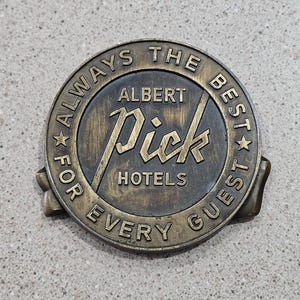 May include: A vintage bronze-colored hotel badge with the text "ALWAYS THE BEST FOR EVERY GUEST" encircling the words "ALBERT Pick HOTELS". The badge has a circular design with a ribbon-like element at the bottom.